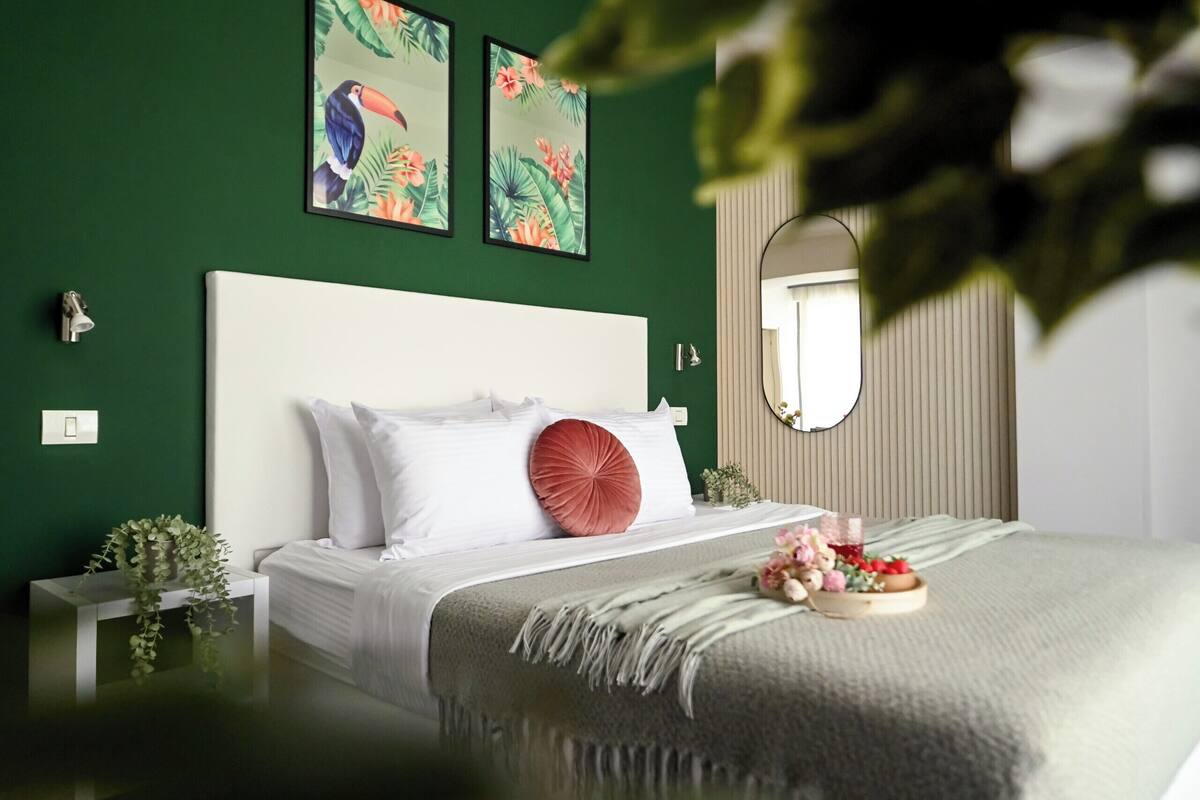 Un dormitorio con una cama, una mesita de noche, un espejo y una pared verde con dos cuadros enmarcados.
