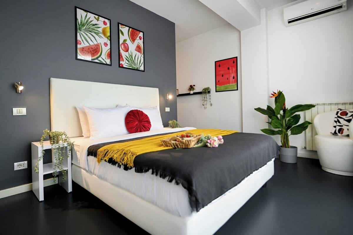 Un dormitorio moderno con una cama grande, una mesita de noche con una planta y una obra de arte en la pared que representa frutas.