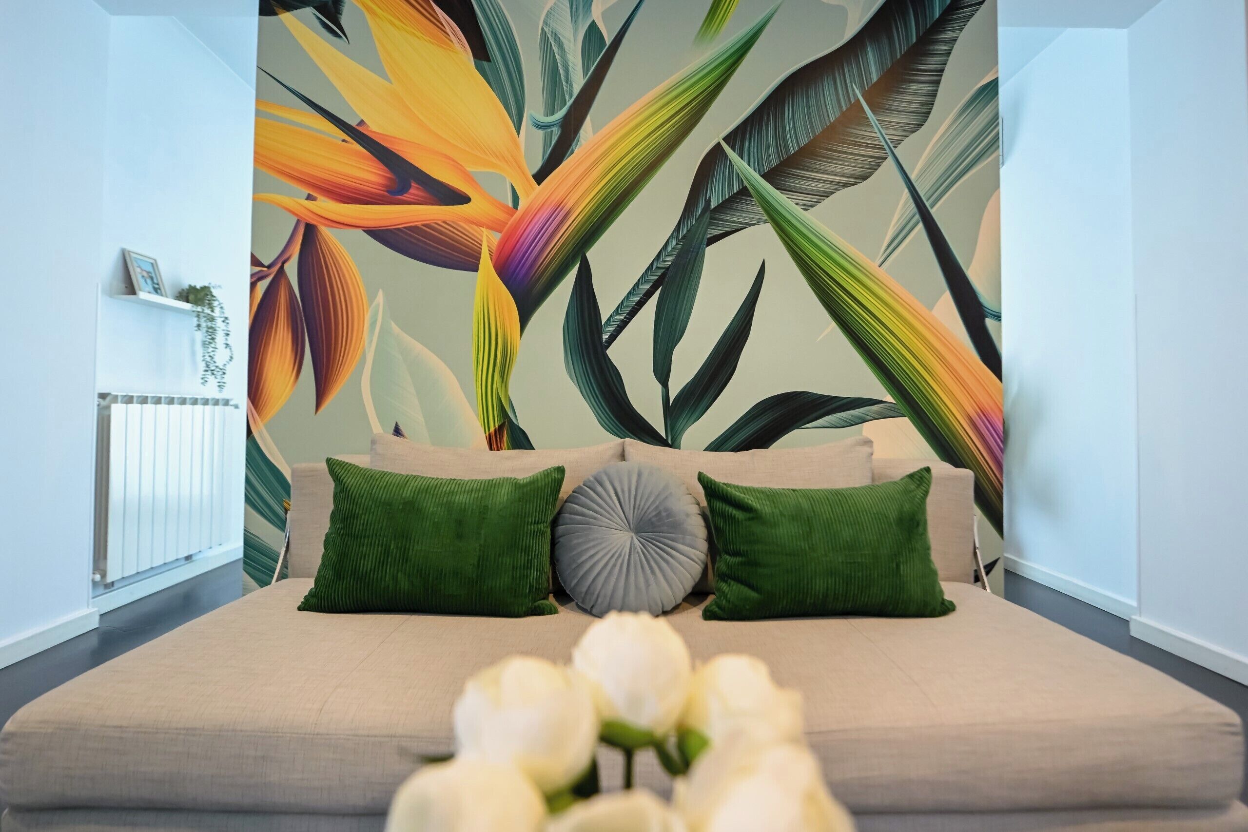 Un dormitorio con una cama, almohadas verdes y grises, un mural floral y un arreglo de flores blancas.