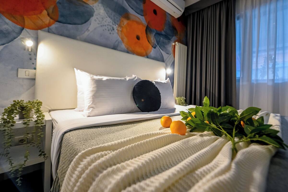 Una cama bien hecha con una colcha blanca, un almohadón oscuro y un almohadón blanco. Hay dos naranjas y una planta verde sobre la cama. La habitación tiene un techo estampado y una ventana con cortinas.