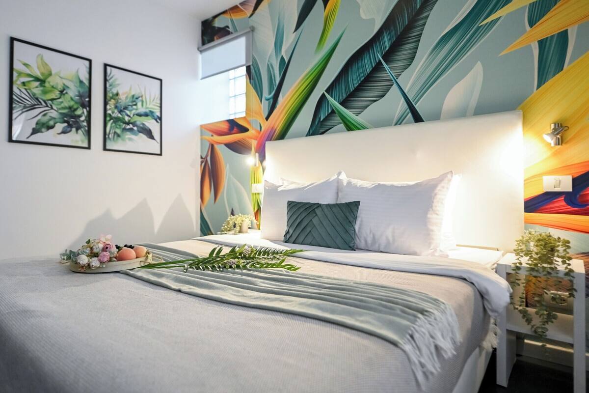Una cama con cabecera blanca, manta gris y almohada verde. Hay dos láminas botánicas enmarcadas en la pared, una planta pequeña en la mesita de noche y un mural con motivos de hojas.