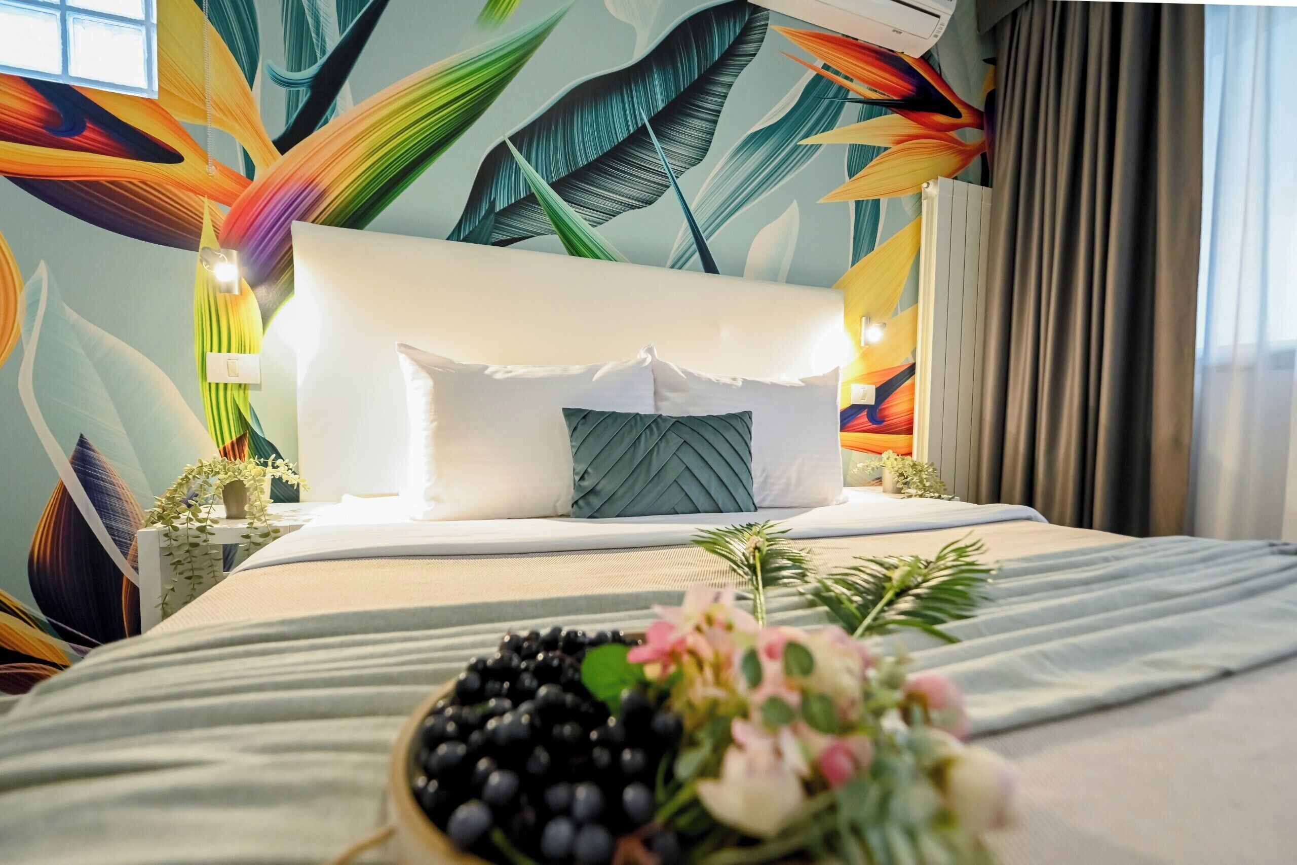 A hotel room with a bed, a headboard, a nightstand, a window, and a wall with a leaf pattern.
