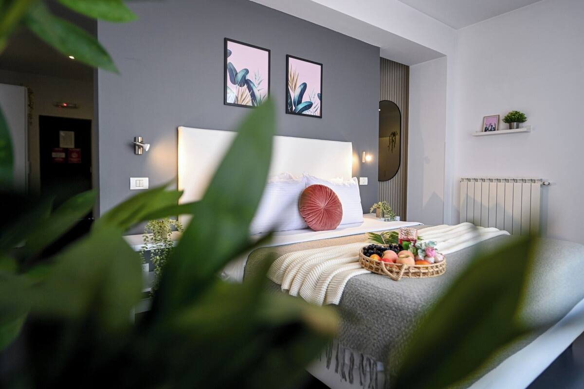 Un dormitorio moderno con una cama grande, una mesita de noche con una cesta de frutas, un espejo montado en la pared y dos obras de arte enmarcadas.