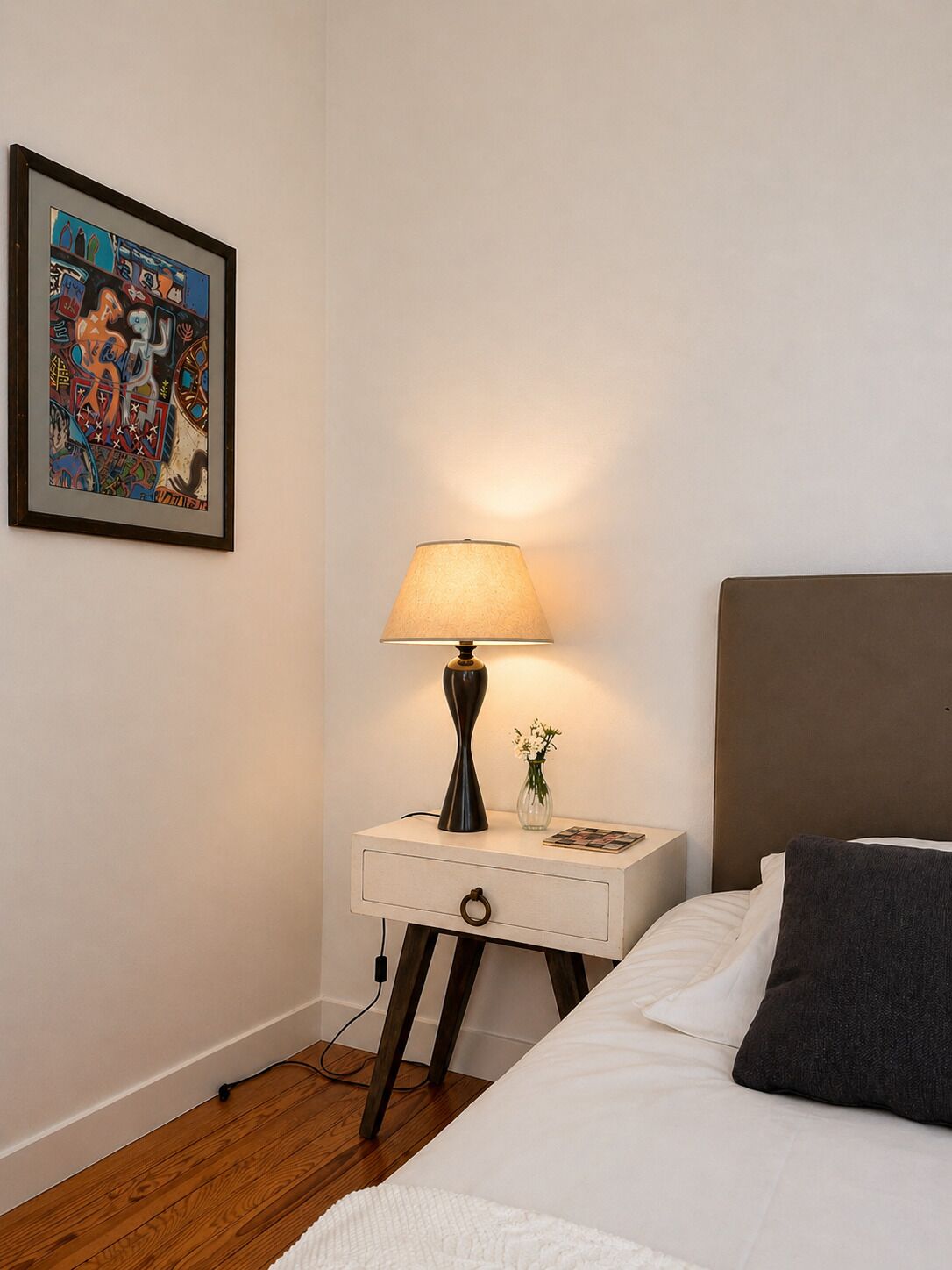 A neatly arranged bedroom with a bed, a bedside table with a lamp, a framed picture on the wall, and a small vase with flowers.