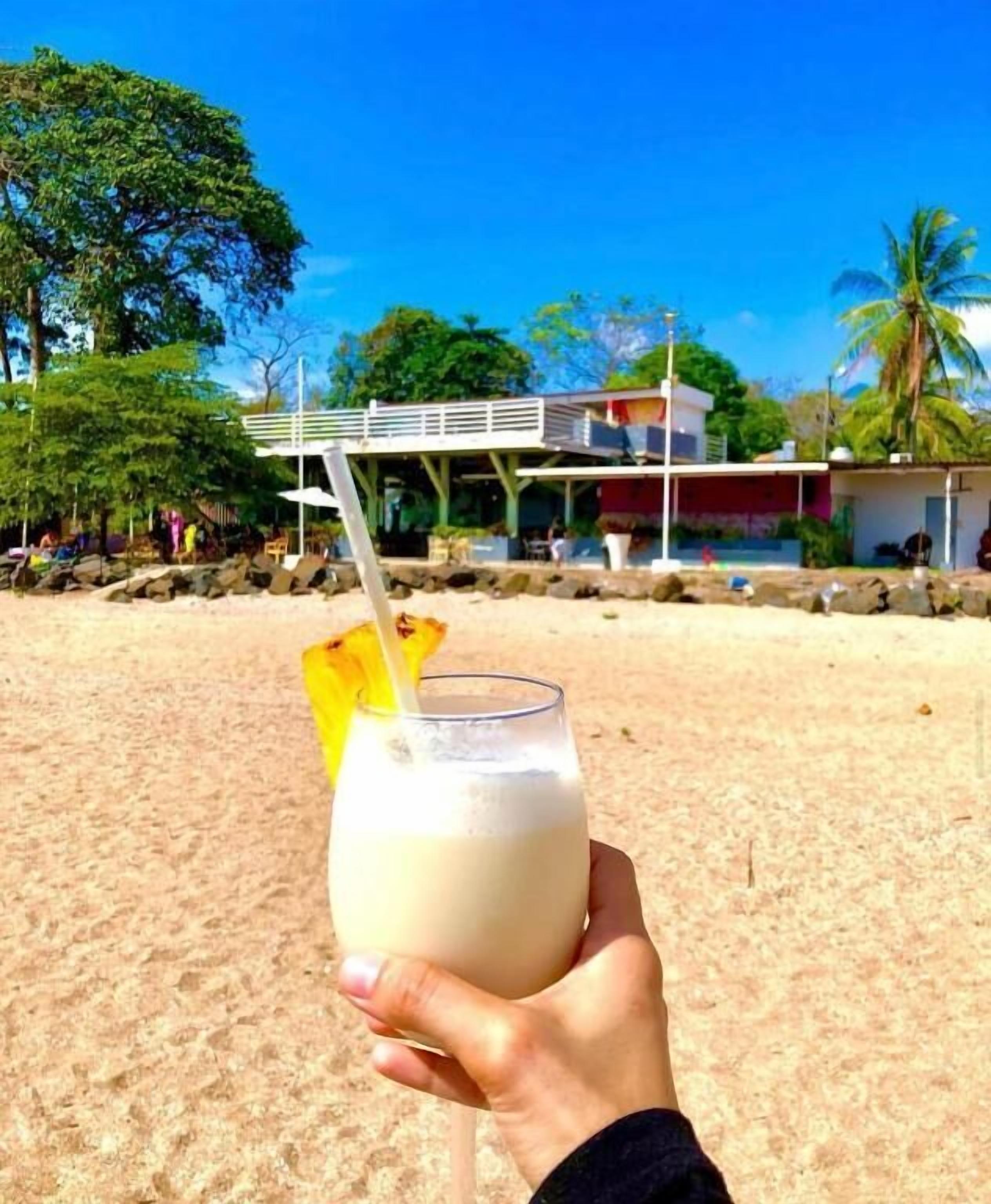 Une main tenant un verre avec une paille et une tranche de fruit sur une plage de sable, avec un bâtiment en arrière-plan.
