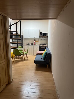 Una cocina compacta con armarios blancos, un microondas y una estufa. Hay una zona de comedor con una silla verde y una mesita. Un sofá con cojines se encuentra en la esquina.