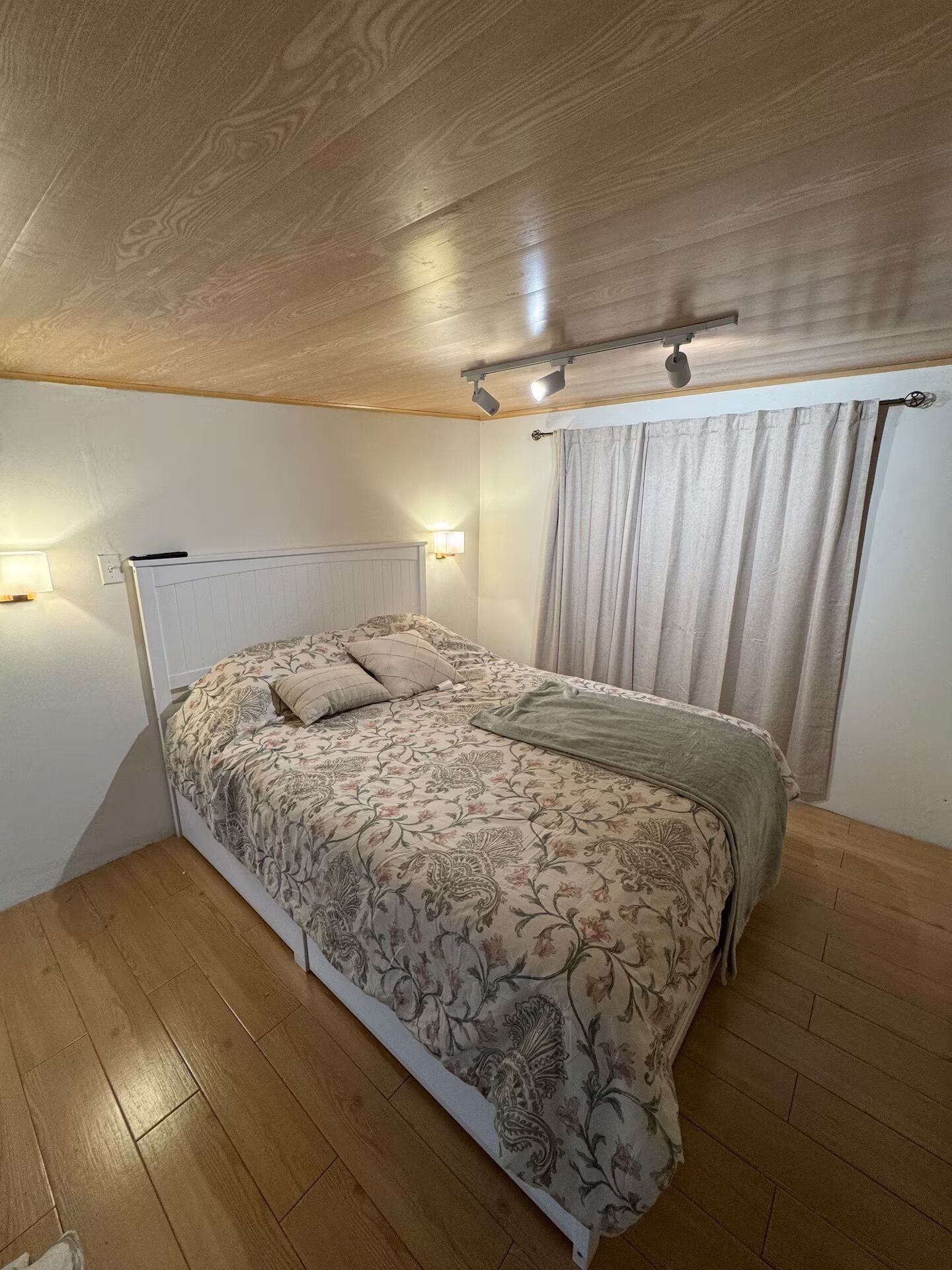 A bedroom with a wooden ceiling, a bed with floral bedding, two wall-mounted lights, and a window with curtains.