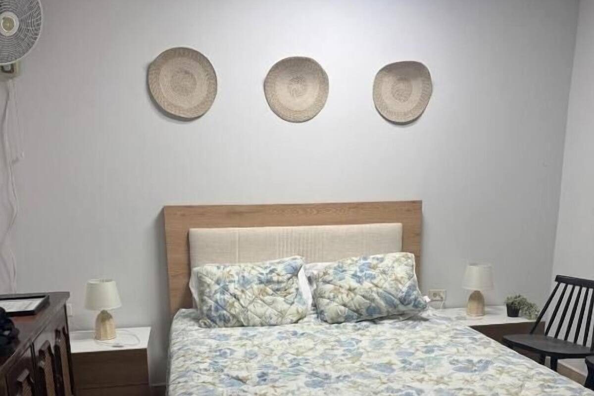 A bedroom with a wooden bed, a bedside table, a lamp, and a wall-mounted fan.