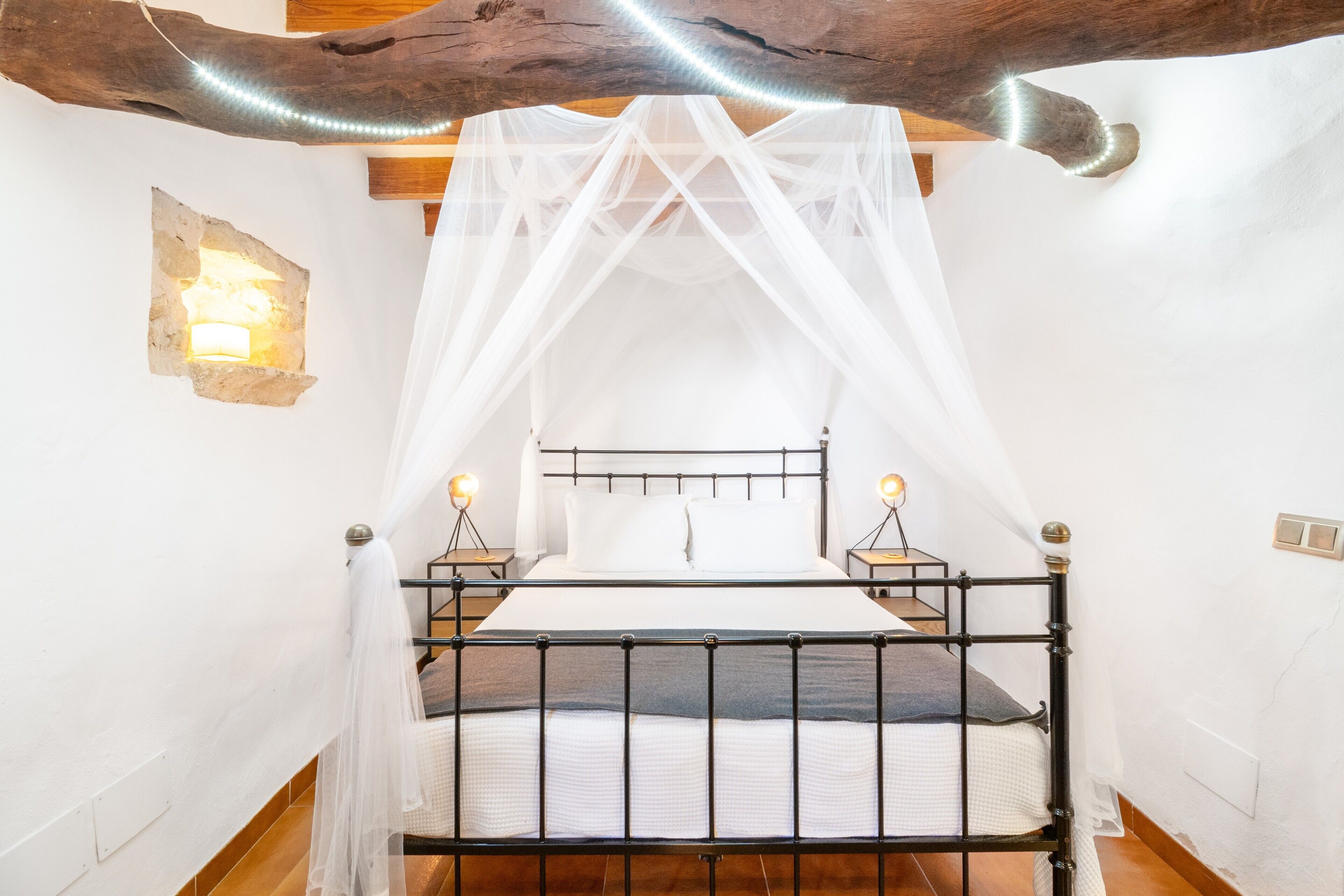 Een bed met een metalen frame, witte beddengoed en een hemelbed. Twee bedlampen en een wandlamp zijn aanwezig.