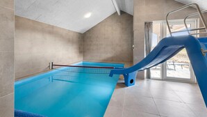 Piscina interior con un tobogán azul, paredes de azulejos y una entrada de cristal transparente.