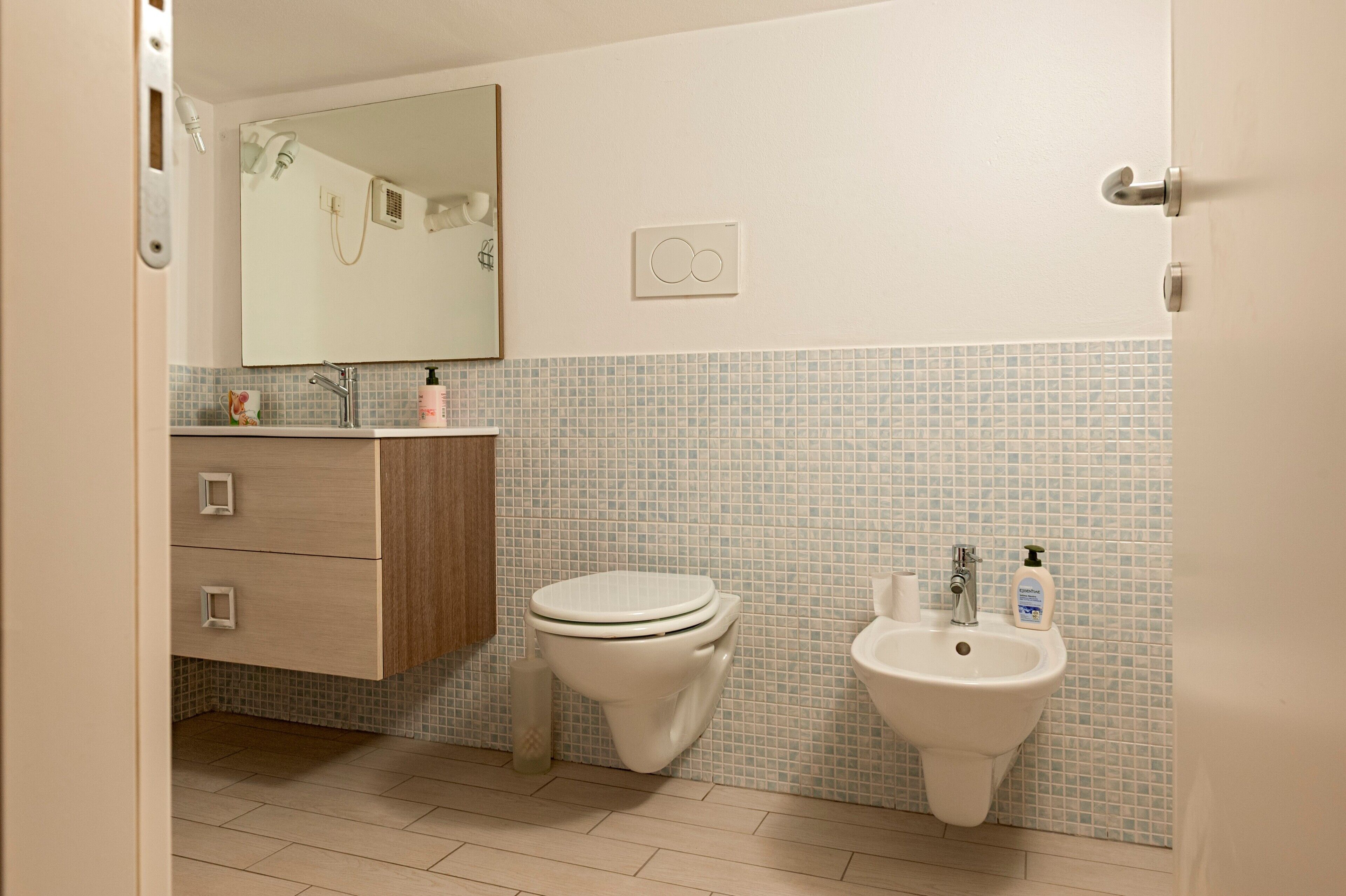 Une salle de bain équipée de toilettes, d’un lavabo, d’un miroir et d’un mur recouvert de carreaux.