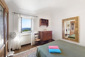 Une chambre à coucher comprenant un lit, une commode, une chaise, un ventilateur, une fenêtre avec des rideaux, un miroir et une vue sur l’extérieur.