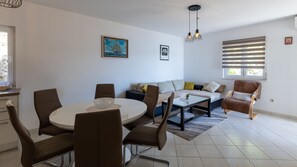 A modern living room with a dining table, chairs, a sofa, and a window with blinds. - Apartman Jelinic 2 by Villas Guide (Klek)