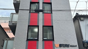 A multi-story building with a prominent red and grey facade, a visible storefront with a sign, and a vending machine at the ground level. - Ustay nippori (Tokyo)