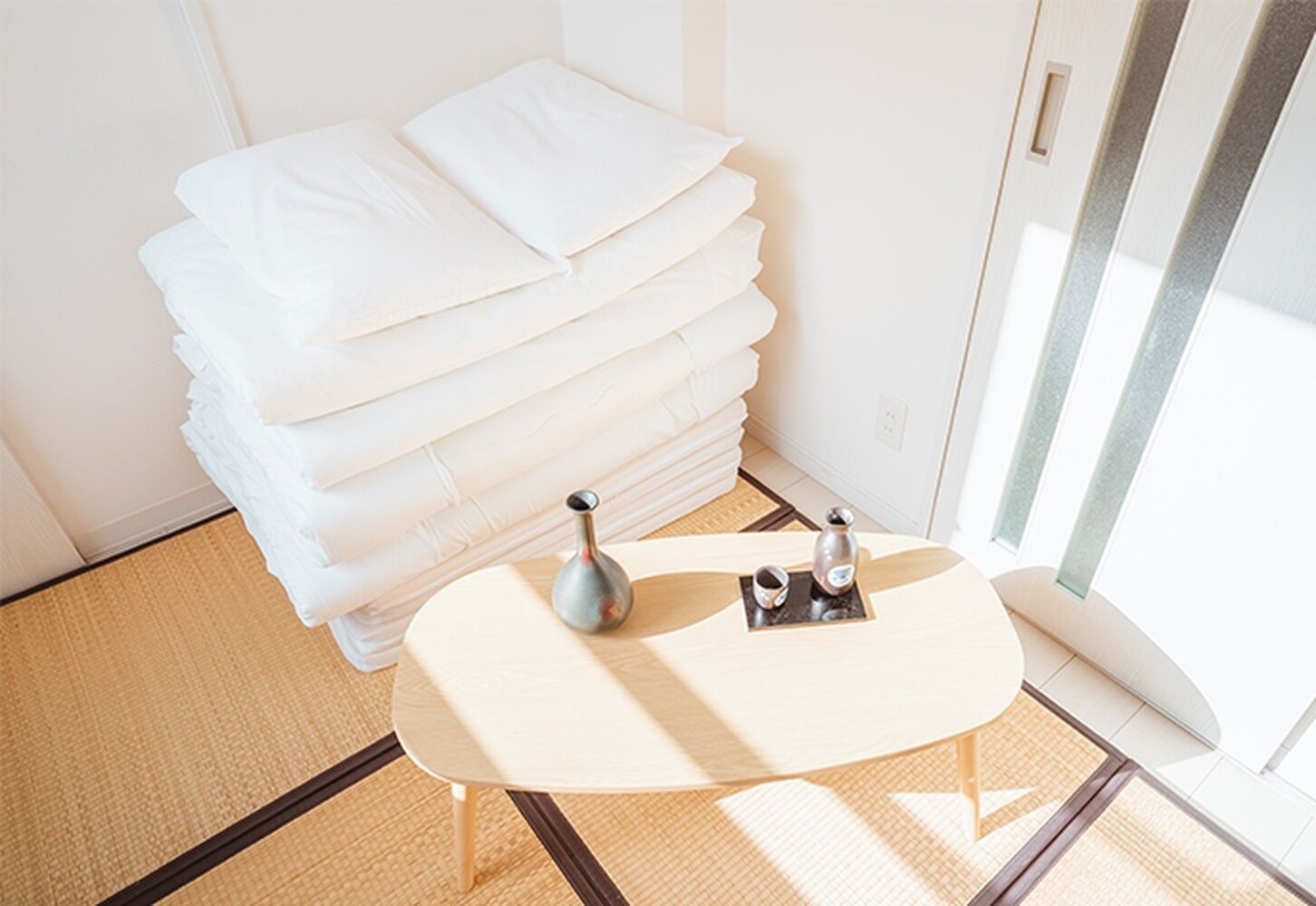 A neatly arranged stack of white pillows in a corner of a room with a small wooden table and two bottles, possibly containing toiletries, on a tray.