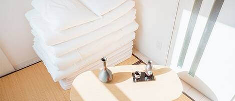 A neatly arranged stack of white pillows in a corner of a room with a small wooden table and two bottles, possibly containing toiletries, on a tray.