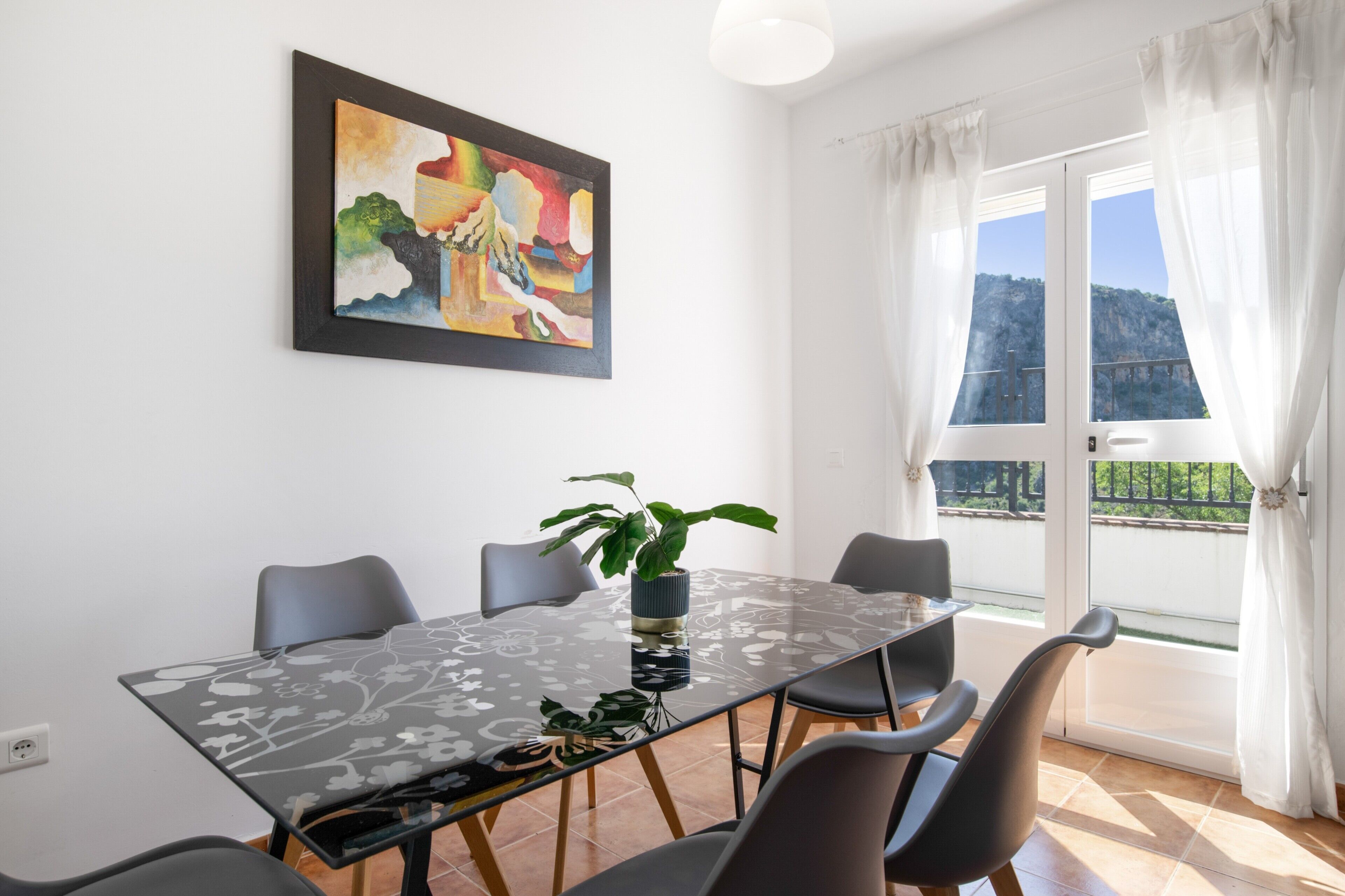 Een eetgedeelte met een glazen tafel, stoelen, een schilderij en uitzicht door een raam.