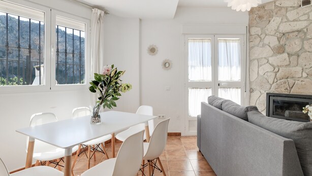 Un coin repas avec une table et des chaises blanches, un canapé gris et un mur en pierre.