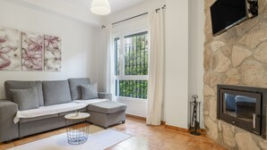 Un saló amb un sofà gris, una taula rodona petita amb una planta, una llar de pedra i un finestral gran amb cortines.