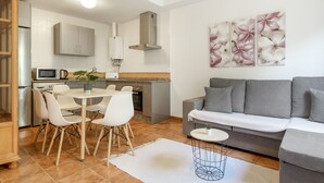 Un espai modern d'estar amb un sofà gris, caires blanques per a dinar, una taula rodona petita i una zona de cuina amb armaris i microones.