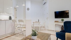 Ett modernt kök med vita skåp, en matplats med vita stolar, en blå soffa och en platt-TV som är monterad på väggen.