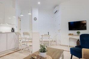 Eine moderne Küche mit weißen Schränken, ein Essbereich mit weißen Stühlen, ein blaues Sofa und ein flachbildfernseher an der Wand.