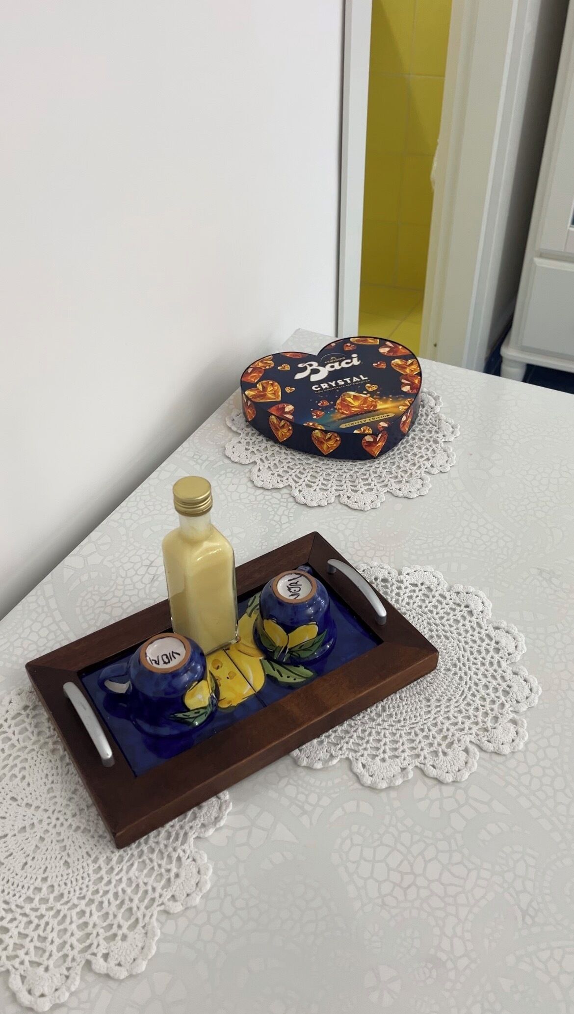 Una bandeja con una botella, una taza y una tetera sobre una mesa con un mantel de encaje. En el fondo, una caja con forma de corazón y diseño floral.