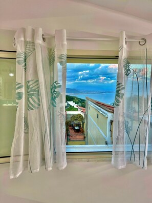 Ventana con cortinas blancas con motivos de hojas, vista de un paisaje urbano costero y balcón con mesa y sillas.