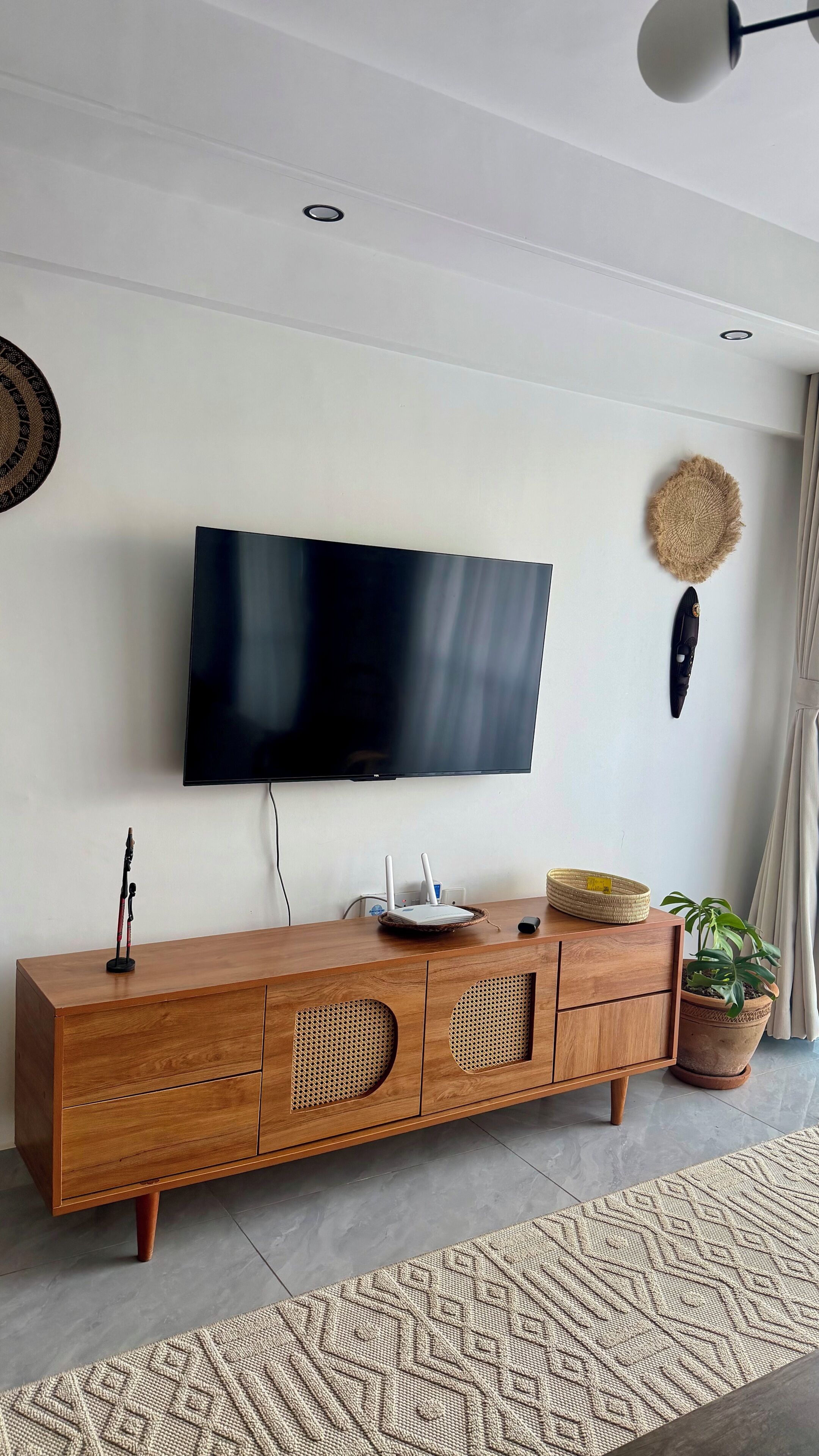 A modern living room with a flat-screen TV mounted on a white wall, a wooden TV stand with woven storage compartments, a woven rug, and a potted plant.