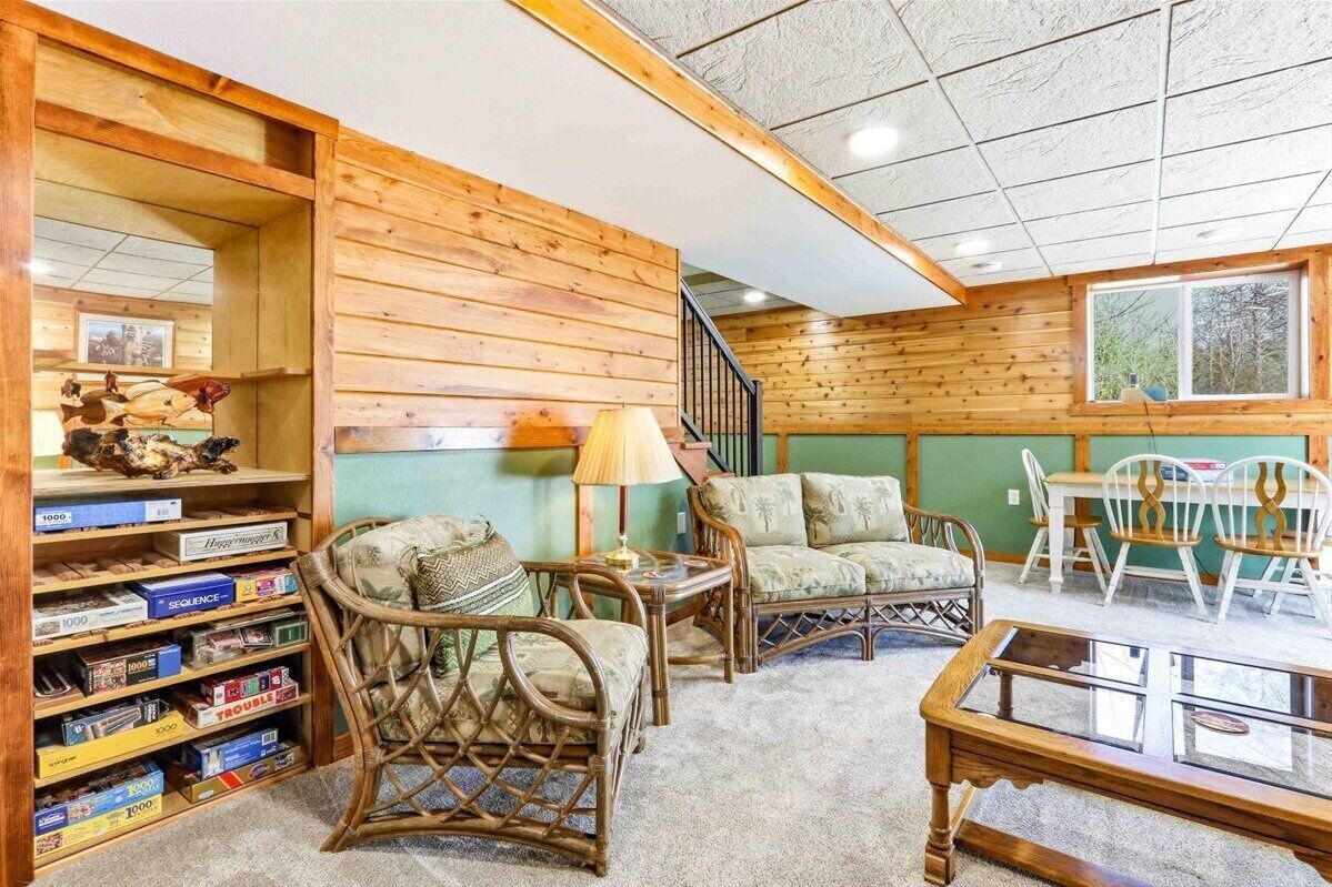 Une pièce chaleureuse aux murs en bois, avec une bibliothèque, des fauteuils en osier, une table en verre et un escalier.
