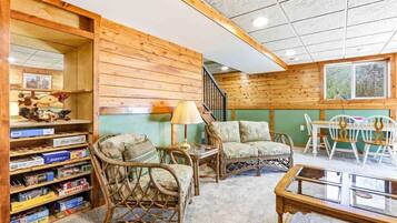 Une pièce chaleureuse aux murs en bois, avec une bibliothèque, des fauteuils en osier, une table en verre et un escalier.