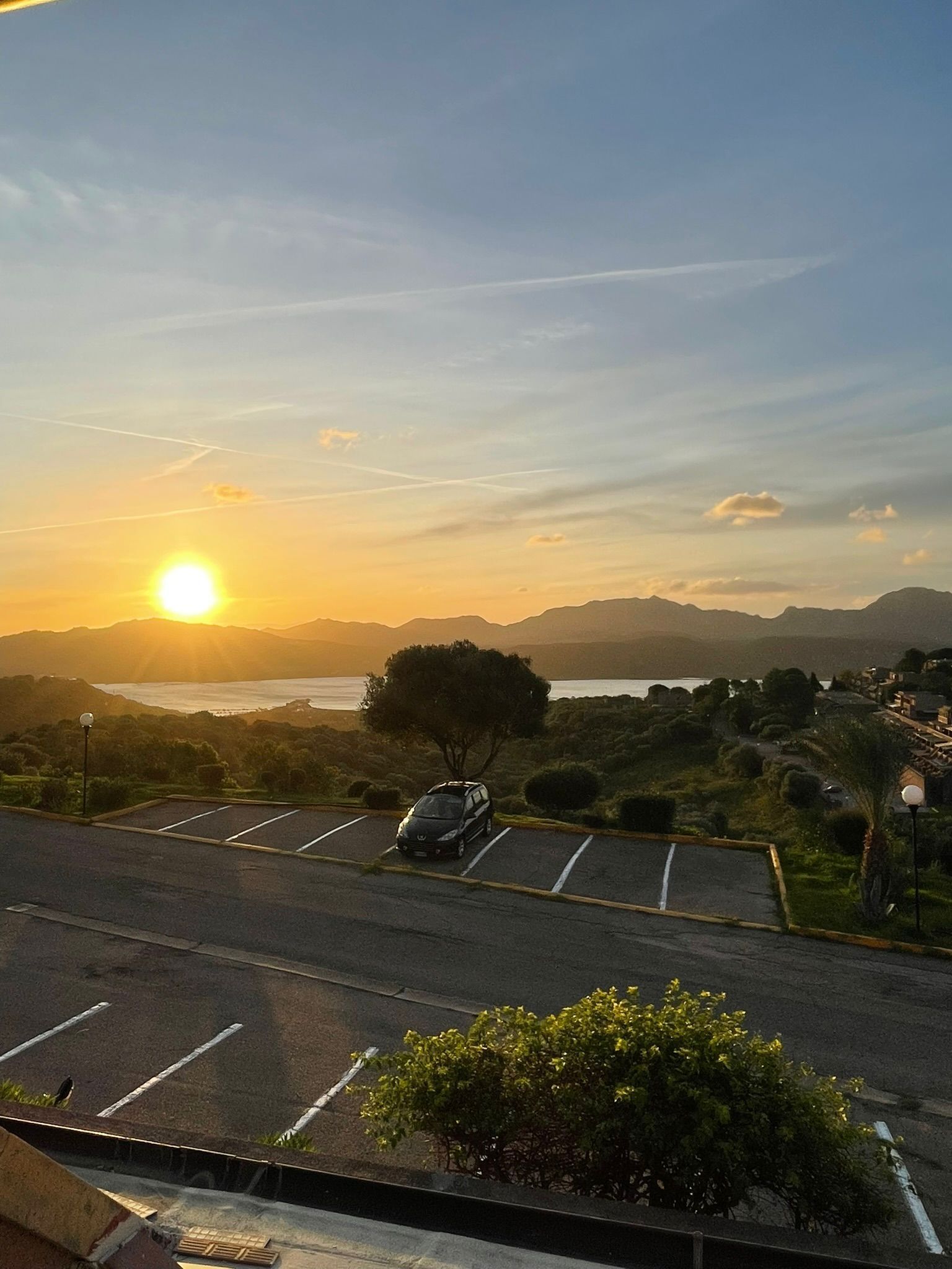 Un parking avec une voiture, un arbre et une vue sur le coucher de soleil au-dessus d’une étendue d’eau et de montagnes.