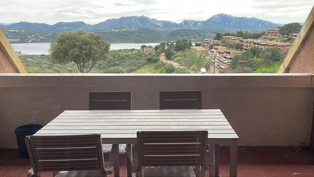 Een balkon met een tafel en stoelen, met uitzicht op een bergachtig landschap en een wateroppervlak.