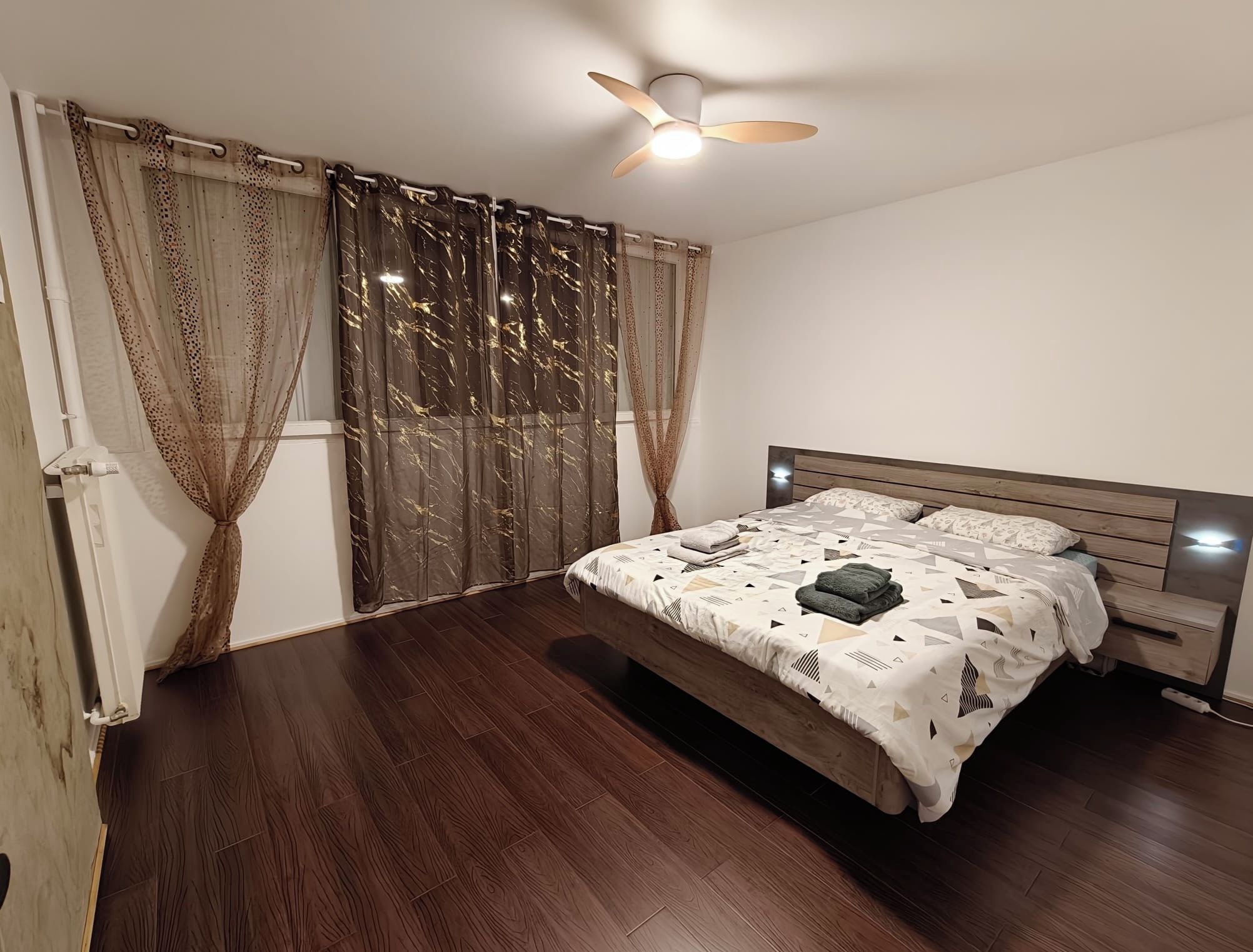 Un dormitorio con una cama de madera, un ventilador de techo y un ventanal con cortinas.