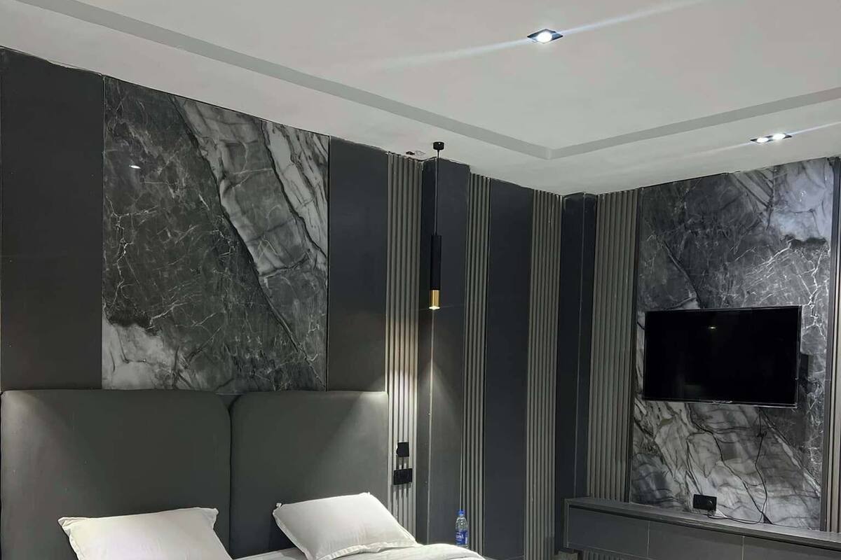 A modern bedroom with a large bed, a flat-screen TV mounted on the wall, and a marble-patterned wall.