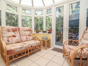 A conservatory with a wicker sofa, a small table, and potted plants. - Honey Cottage (Ilfracombe)