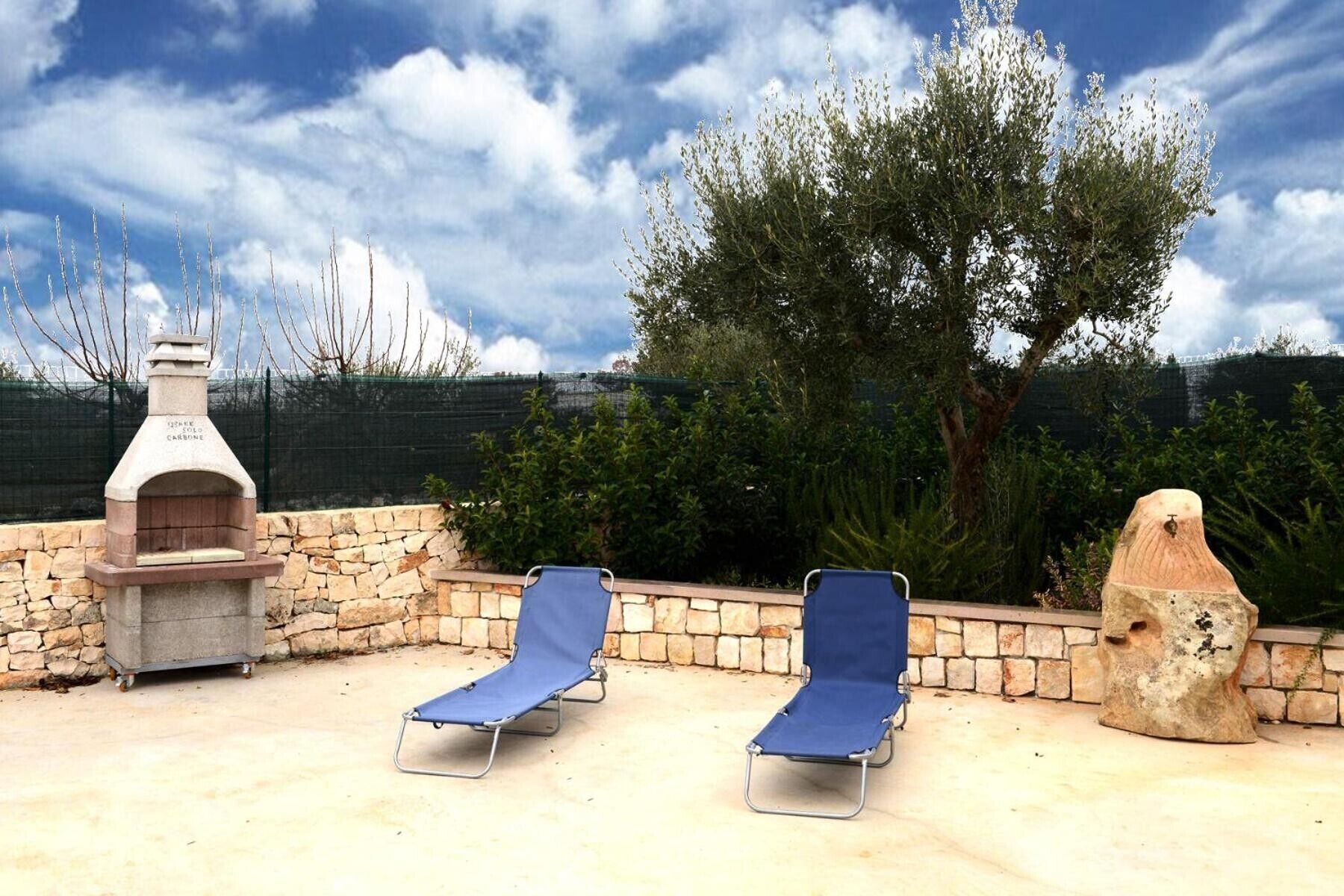 A stone barbecue grill, two blue lounge chairs, and a large tree in a backyard setting.