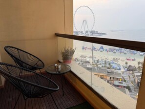 Un balcon avec deux chaises noires, une petite table, un pot de fleurs et une vue sur une plage où se trouve une grande roue.