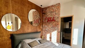 Ein Schlafzimmer mit Backsteinwand, einem runden Spiegel, einem Bett mit grauem Kopfteil und einer zur Garderobe führenden Tür.