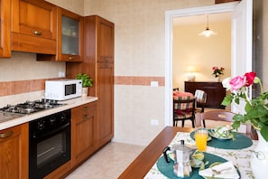 A kitchen with wooden cabinets, a microwave, a stove, and a dining area with a table set for breakfast. - De Vivo Realty - Villa Teresa (Piano di Sorrento)