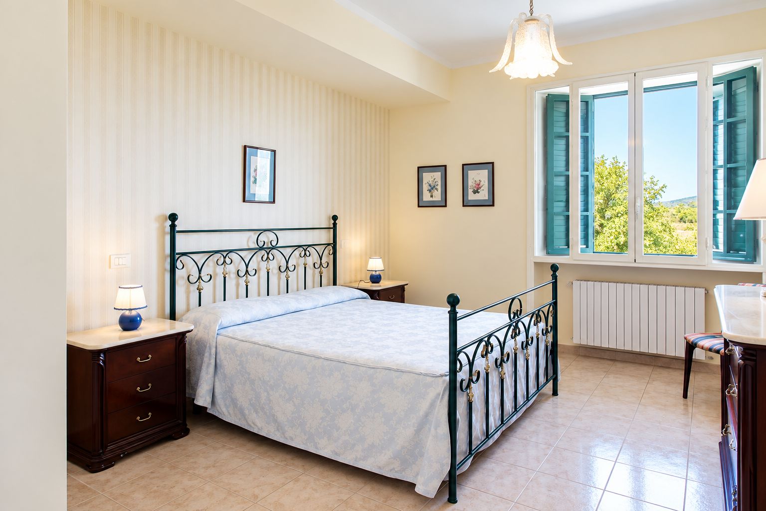 Una camera da letto con un letto, comodini, una cassettiera, una finestra con persiane e un termosifone.