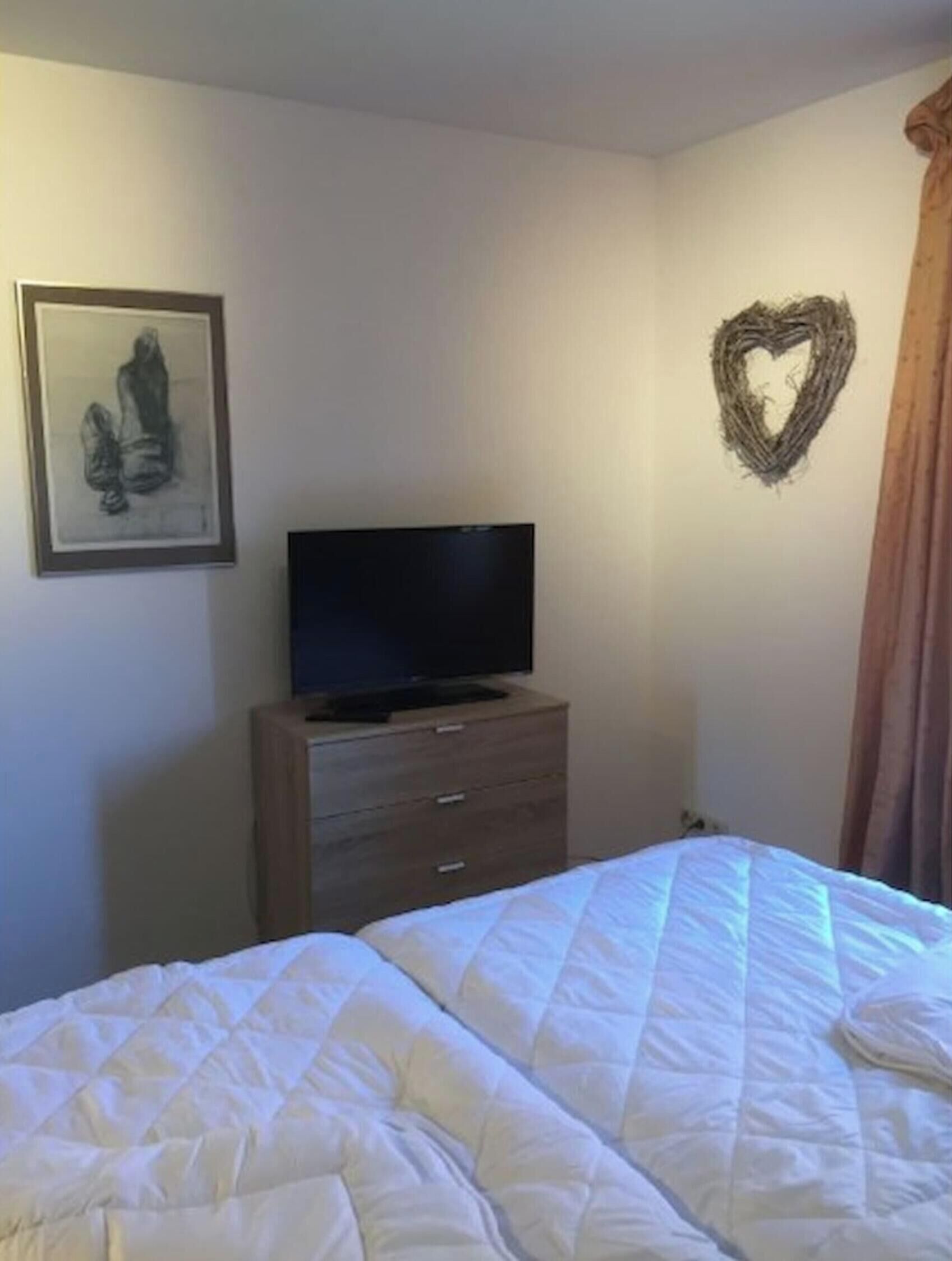 Ein Schlafzimmer mit einem Bett, einem Schminktisch, einem Fernseher, einem gerahmten Bild und einer herzförmigen Dekoration an der Wand.