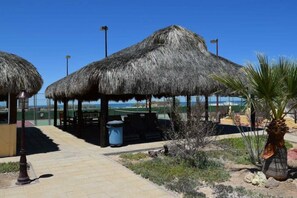 A covered outdoor area with thatched umbrellas, a trash bin, and a palm plant. - Mision Los Sahuaros #72 - 2BR with Rooftop Deck (San Felipe)