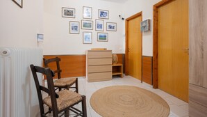 A room with a wooden wardrobe, a radiator, a chair, a small wooden cabinet, and a wall with framed pictures.