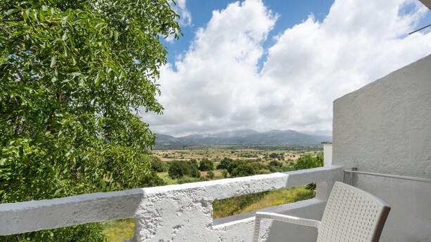 A balcony with a white railing, a wicker chair, and a view of a landscape with mountains and trees.