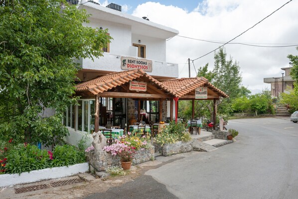 A building with a sign for 'Rent Rooms Dionysios' and a covered outdoor seating area with tables and chairs.