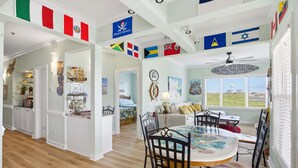 A dining area with a round table and chairs, a ceiling fan, and various flags hanging from the ceiling. - unobstructed ocean views-pool club-private bath for each bedroom-elevator-parking 4 cars-golf cart friendly<br>APRIL '26 UPDATES- new floors, paint, HVAC, renovated walk in shower (Ocean Isle Beach)