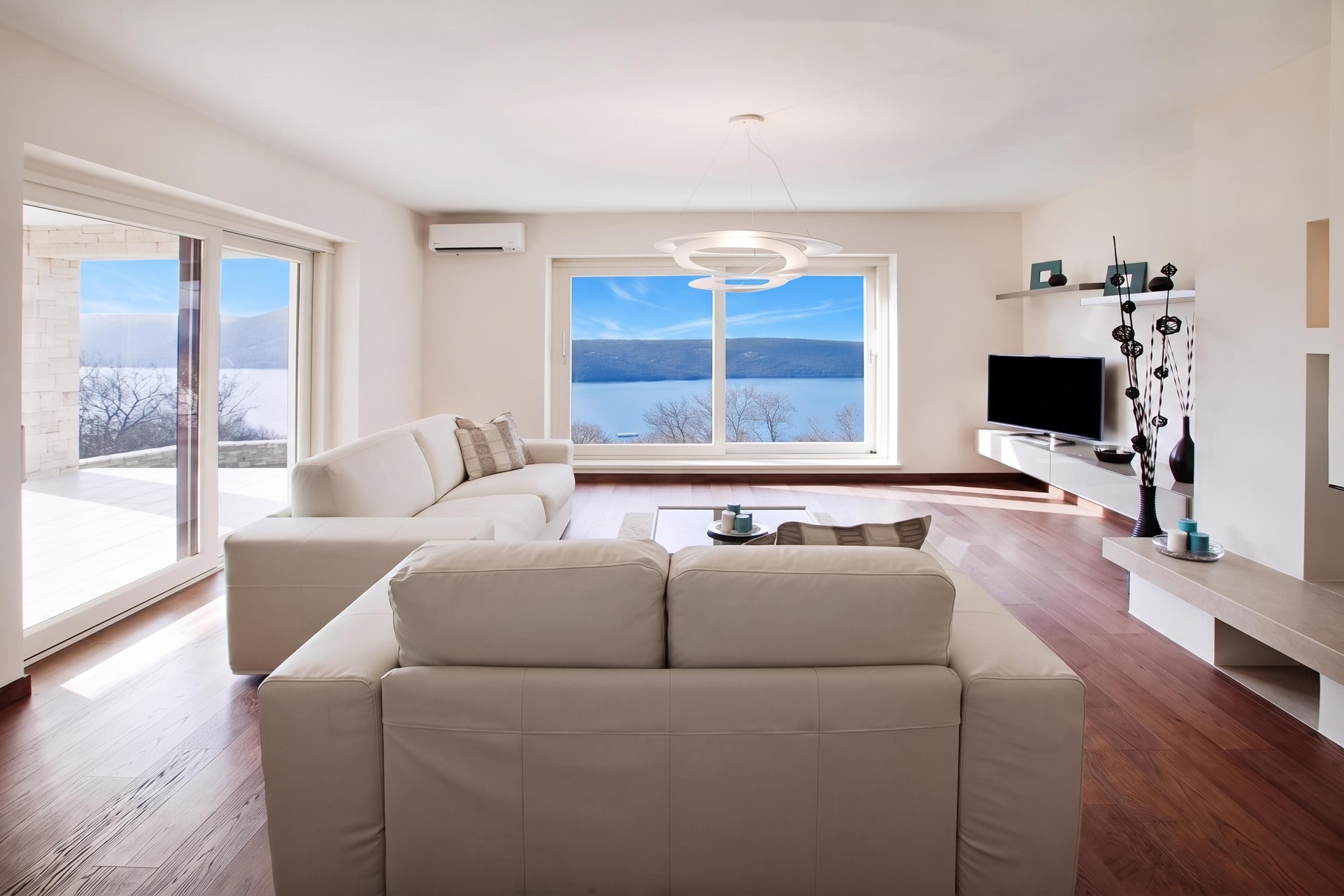 Una sala de estar moderna con un sofá, una mesa de centro y un televisor. Se ve un ventanal con vistas a un lago y montañas.