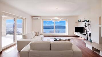 Una sala de estar moderna con un sofá, una mesa de centro y un televisor. Se ve un ventanal con vistas a un lago y montañas.