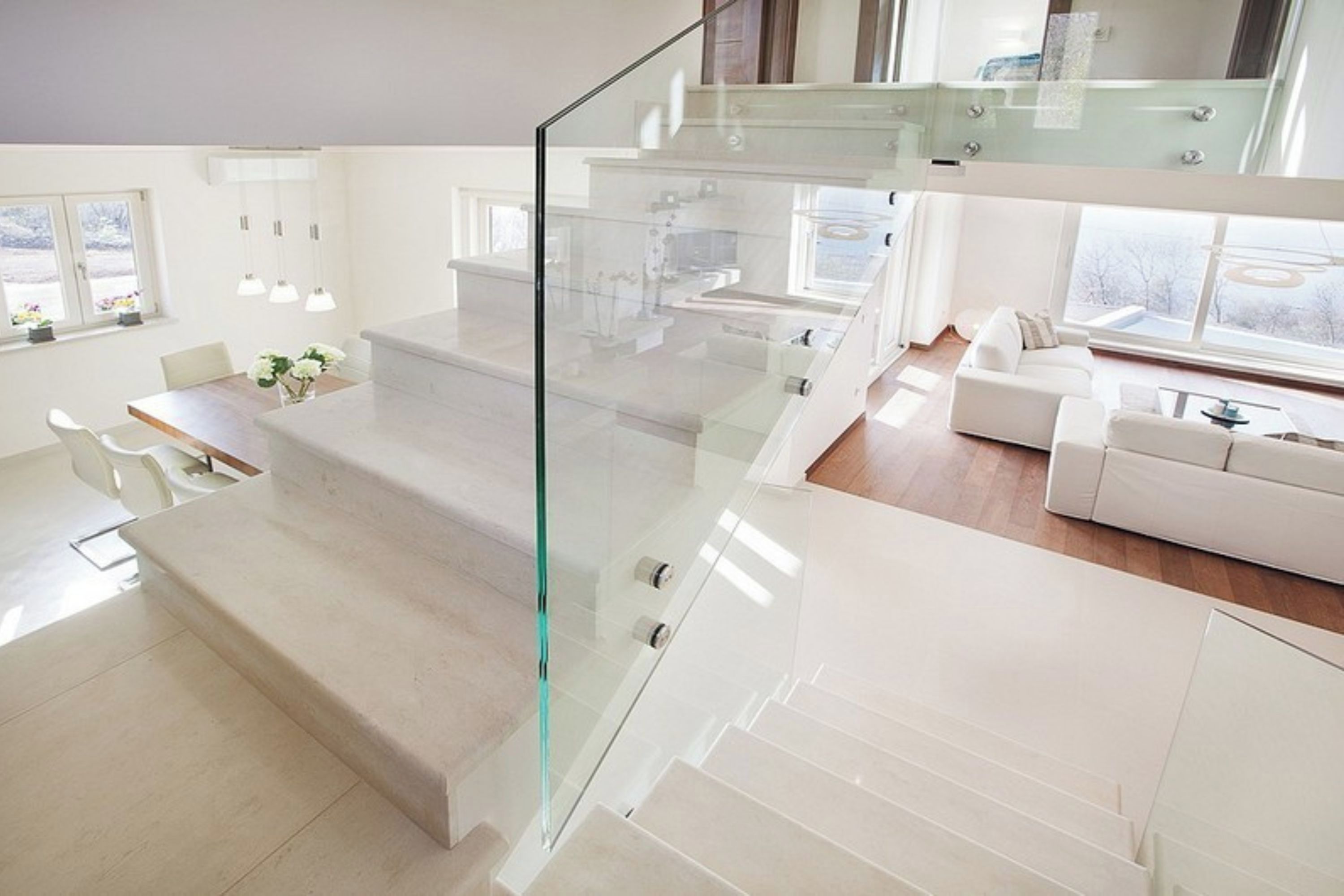 Un interior moderno con una escalera de cristal, zona de comedor, sala de estar y ventanas amplias.